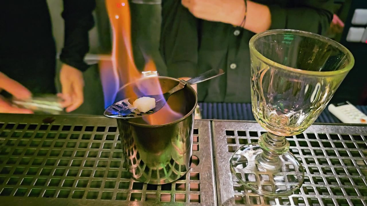 Servizio di cocktail fiammeggiante in un bar poco illuminato, con mani del barista, fiamme blu, shaker in metallo, cucchiaio forato, cubetto di zucchero, calice di vetro e bancone in acciaio inossidabile riflettente