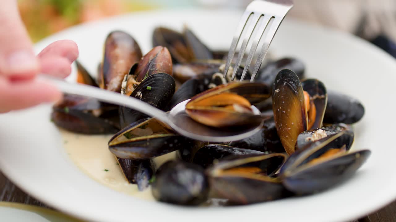 Hand uses fork and spoon to eat steamed mussels on white plate in natural light