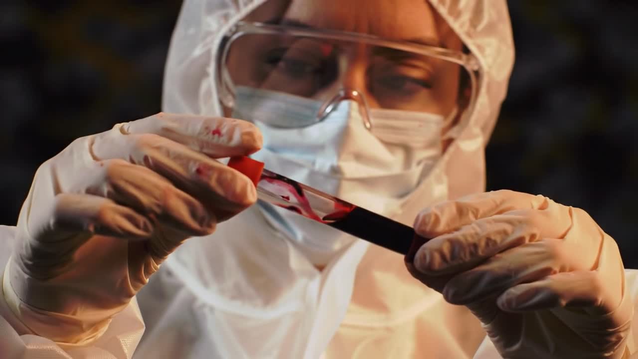 A laboratory worker carefully examines a blood sample in a sterile environment while wearing protective clothing and equipment. The focus is on precision and safety in the analysis process.