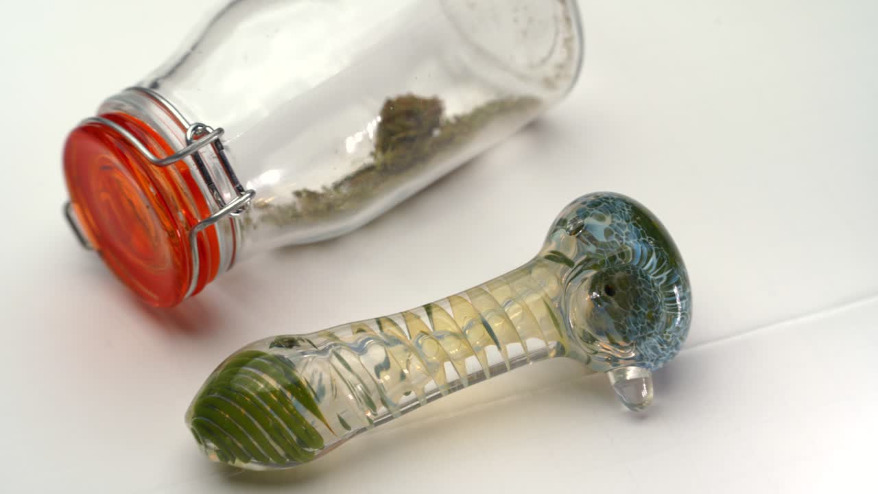 una pipa de vidrio de colores junto a una botella transparente sellada que contiene marihuana