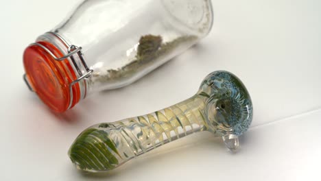 una pipa de vidrio de colores junto a una botella transparente sellada que contiene marihuana