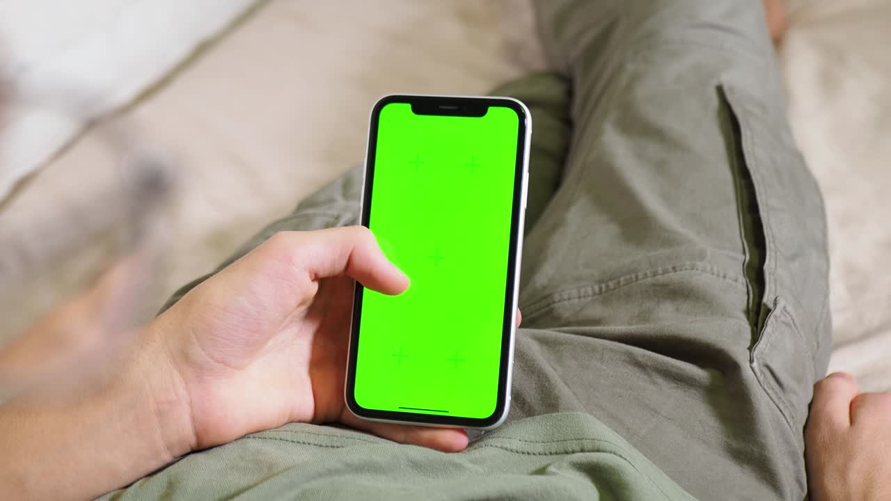 a phone with a green screen in the hands of a man lying on the bed. Modern technologies in the life of adolescents. Phone chroma. Green screen chrom key. watch from behind