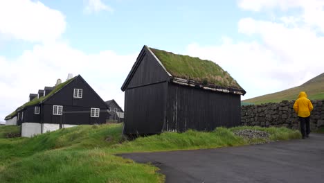 Rückaufnahme-Eines-Mannes-In-Einem-Gelben-Regenmantel,-Der-Auf-Traditionelle-Färöische-Häuser-Mit-Torfdach-In-Vidareidi-Zugeht