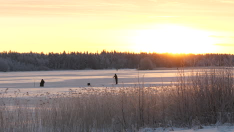 Fisherman-drilling-hole-for-fishing-on-frozen-lake-during-sunset,-Long-distance-shot