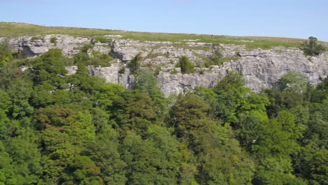 Drone-footage-flying-sideways-and-ascending-up-and-over-a-steep,-tree-lined-limestone-crag-revealing-the-Yorkshire-countryside-with-farmland,-fields,-stone-walls-and-thehilly-landscape-in-the-distance