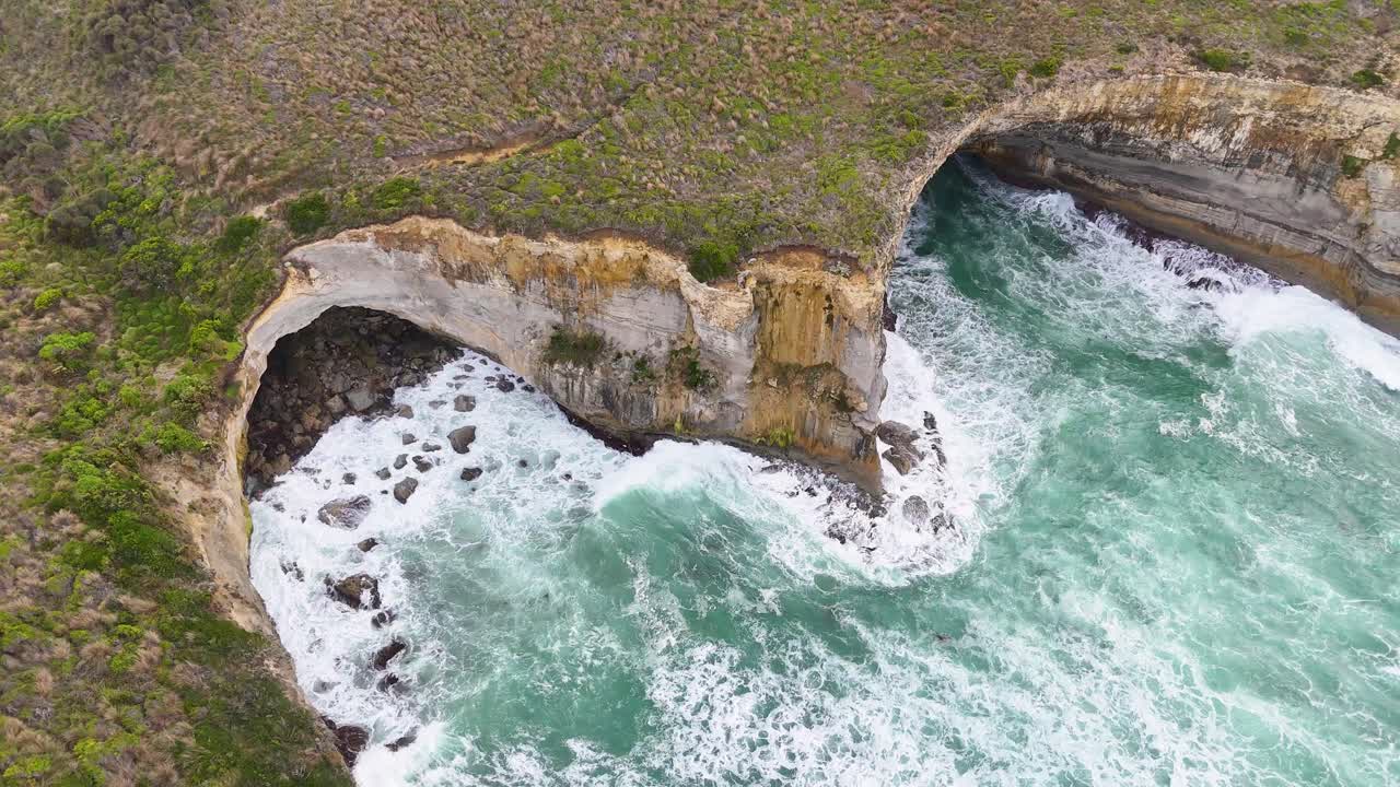 Drone footage captures waves crashing against cliffs at Port Campbell, Australia. Dynamic aerial perspective highlights natural erosion and vibrant ocean hues