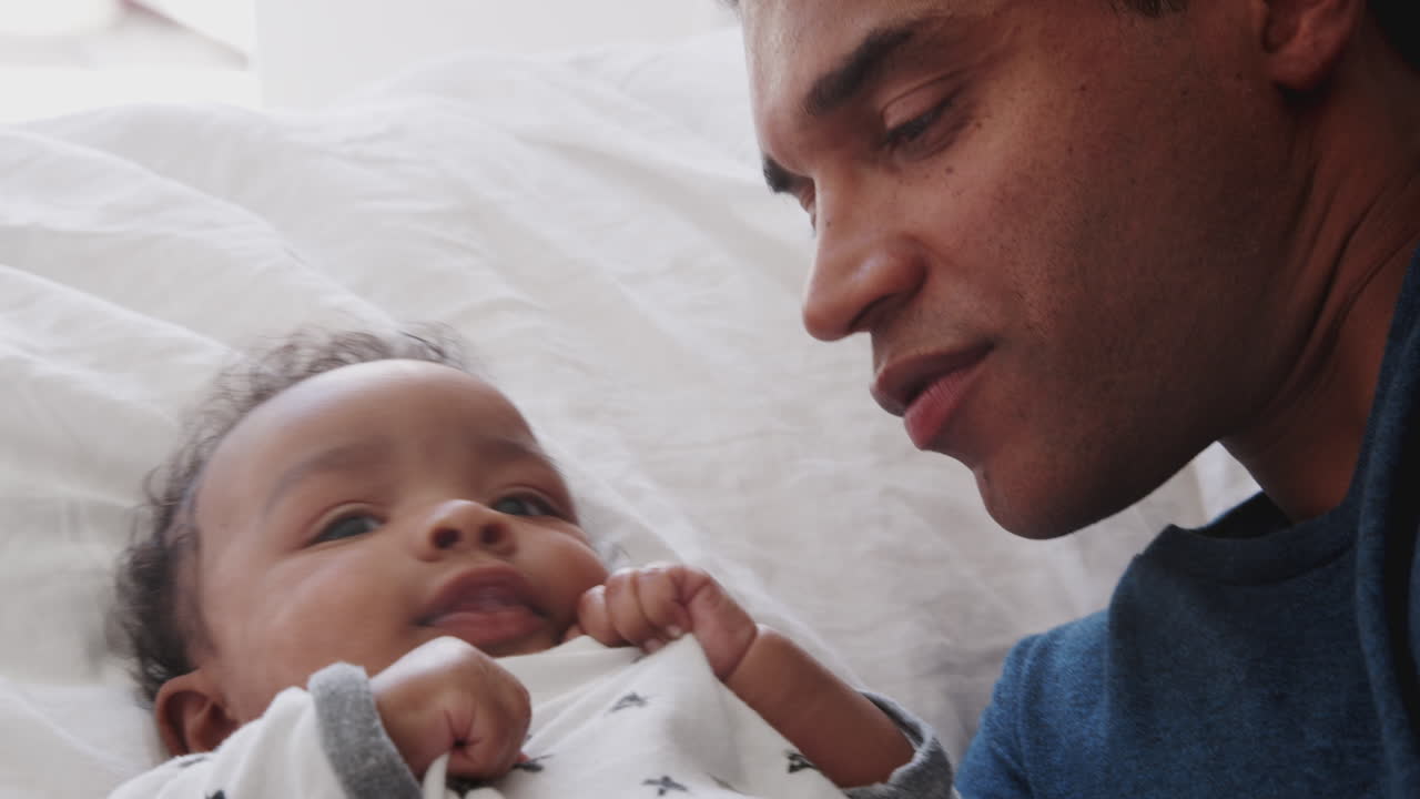 Close of millennial African American father lying on the bed kissing his baby son