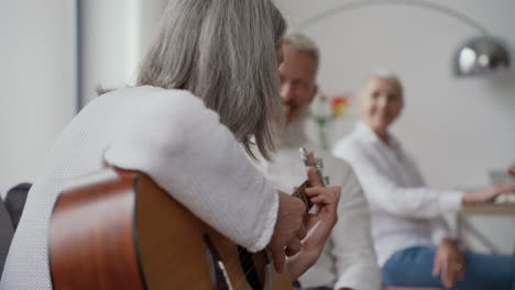 feliz anciana cantando y tocando la guitarra sentada en una silla, mientras en un fondo borroso amigos mayores la escuchan y cantan juntos sentados en la mesa