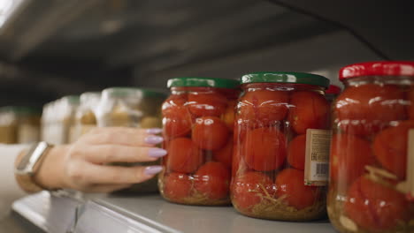 close up of elegant woman touching glass jar of tomatoes on store shelf rails highlighting polished nails wrist watch and blurred aisle background in bright retail setting customer interaction moment