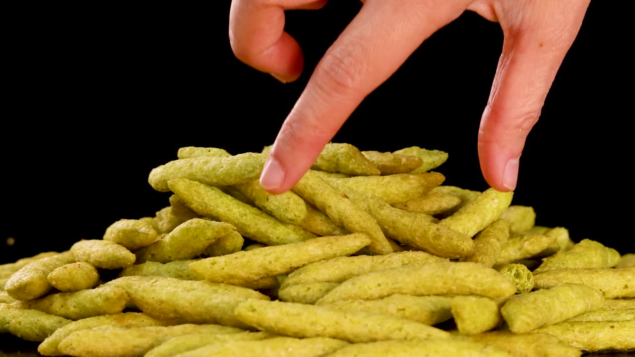 Hand selects green pea puffed snack stick from pile under studio lighting, black background