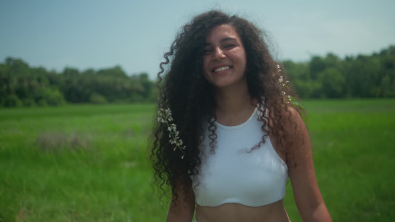 A woman walks with flowers and leaves in her hair, turns to the camera smiling, then walks away in a green field