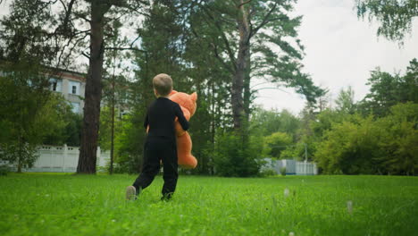 rear view of young boy dressed in black holding large orange teddy bear while walking playfully across grassy park surrounded by trees, with residential buildings partially visible in background