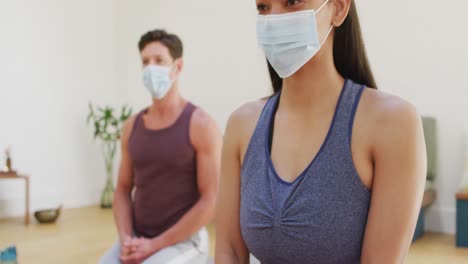 Diverse-man-and-woman-in-face-masks-kneeling-on-mats-at-yoga-class-listening-to-instructor