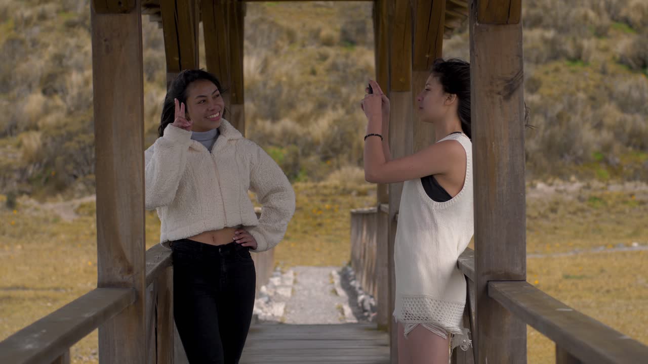Two friends take pictures and pose on wooden bridge on windy day, cotopaxi national park