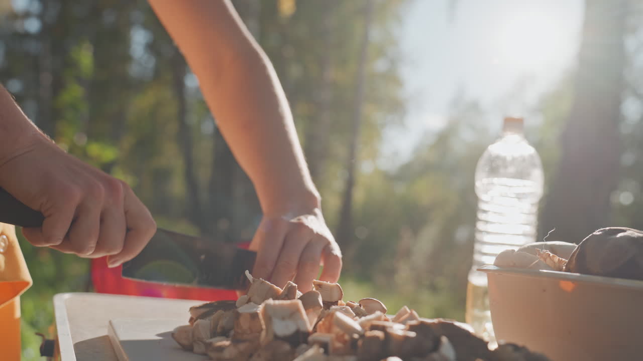 White woman slicing cooked meat campsite cutting board sunlight. Closeup hands guide knife through diced meat, bowl and water bottle visible, red chair background, focused culinary prep for outdoor