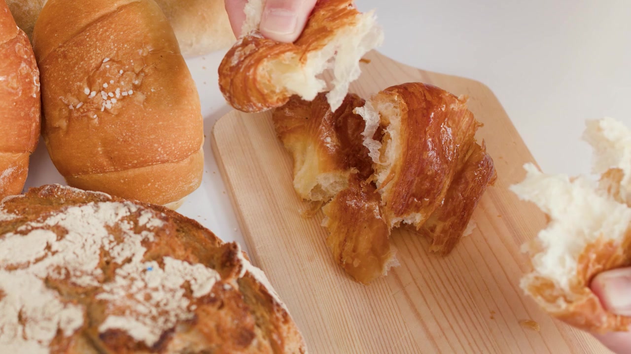 Close-up of hands breaking flaky croissant on wooden board surrounded by sourdough and baguettes.