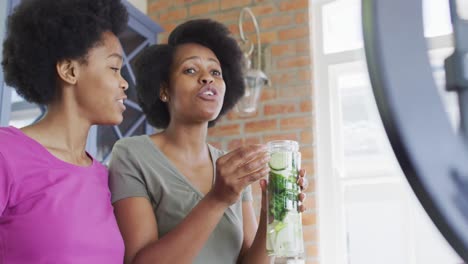 Happy-african-american-mother-and-daughter-preparing-healthy-drink,-making-video-using-smartphone