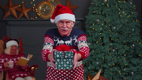 Mature-grandfather-in-New-Year-sweater-and-hat-presenting-Christmas-gift-box,-holidays-at-home