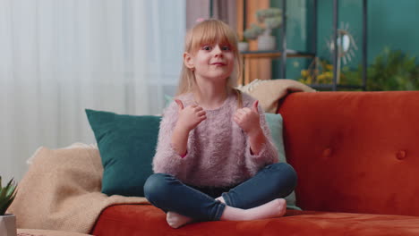 child girl kid sitting on sofa at home alone showing thumbs up like sign positive something good
