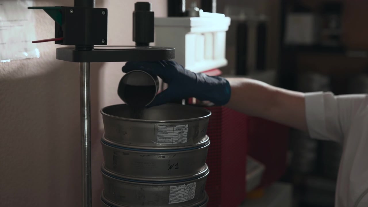 A person wearing a red shirt and gloves is pouring liquid from a bucket into another container in an indoor setting with wooden furniture and shelves. The scene captures a neutral mood.