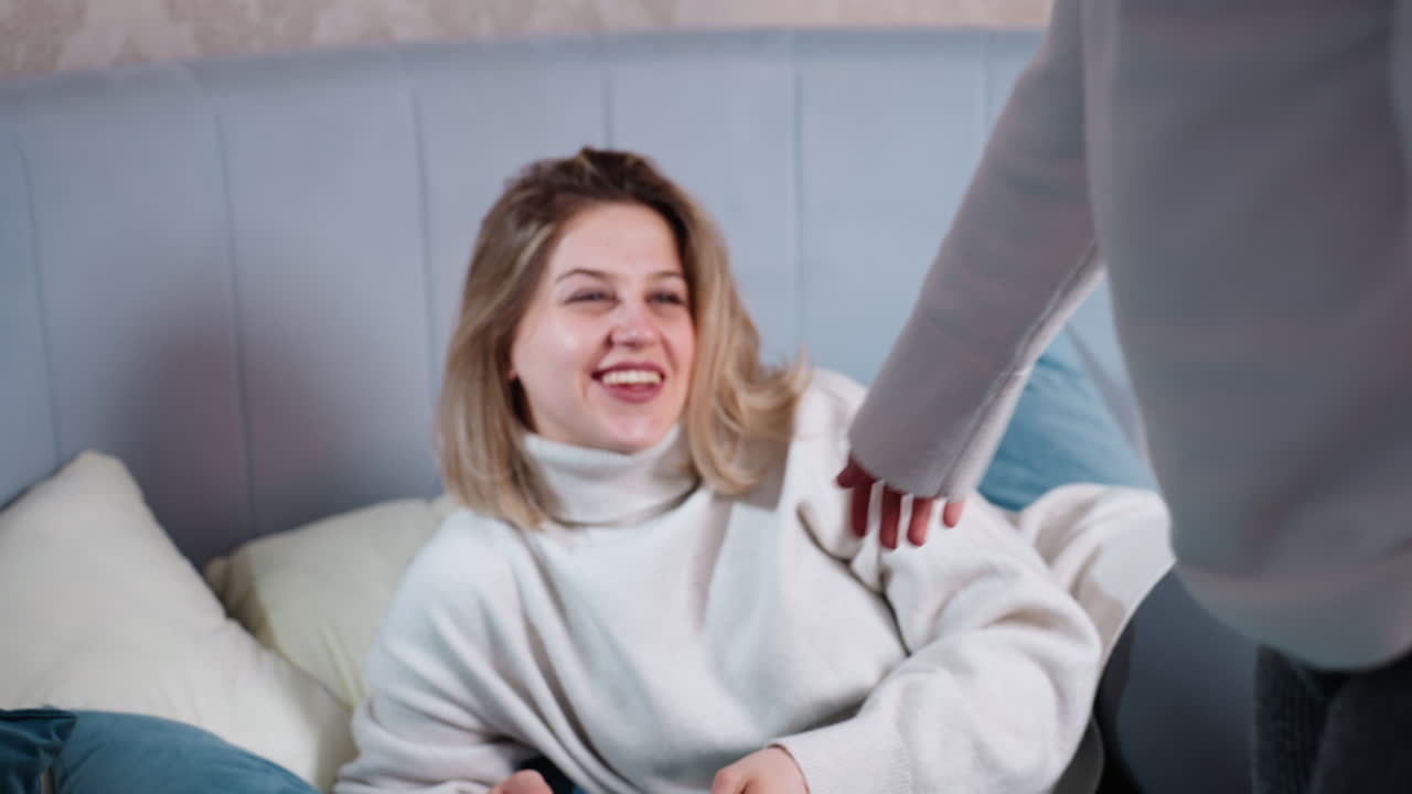 Smiling woman in light sweater sits on bed holding notebook while young girl with ponytails jumps nearby, creating warm family moment full of connection, joy, bonding, and shared activity in cozy home