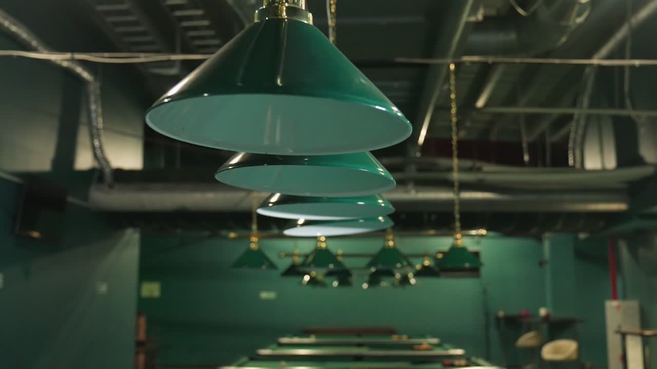 빌리어드 방에서 초록색 색조를 가진 오버헤드 램프의, 황금 금속의 우아한 디자인을 강조합니다. 따뜻한 조명은 클래식한 분위기를 향상시니다. 배경에는 여러 개의 풀 테이블을 반영합니다.