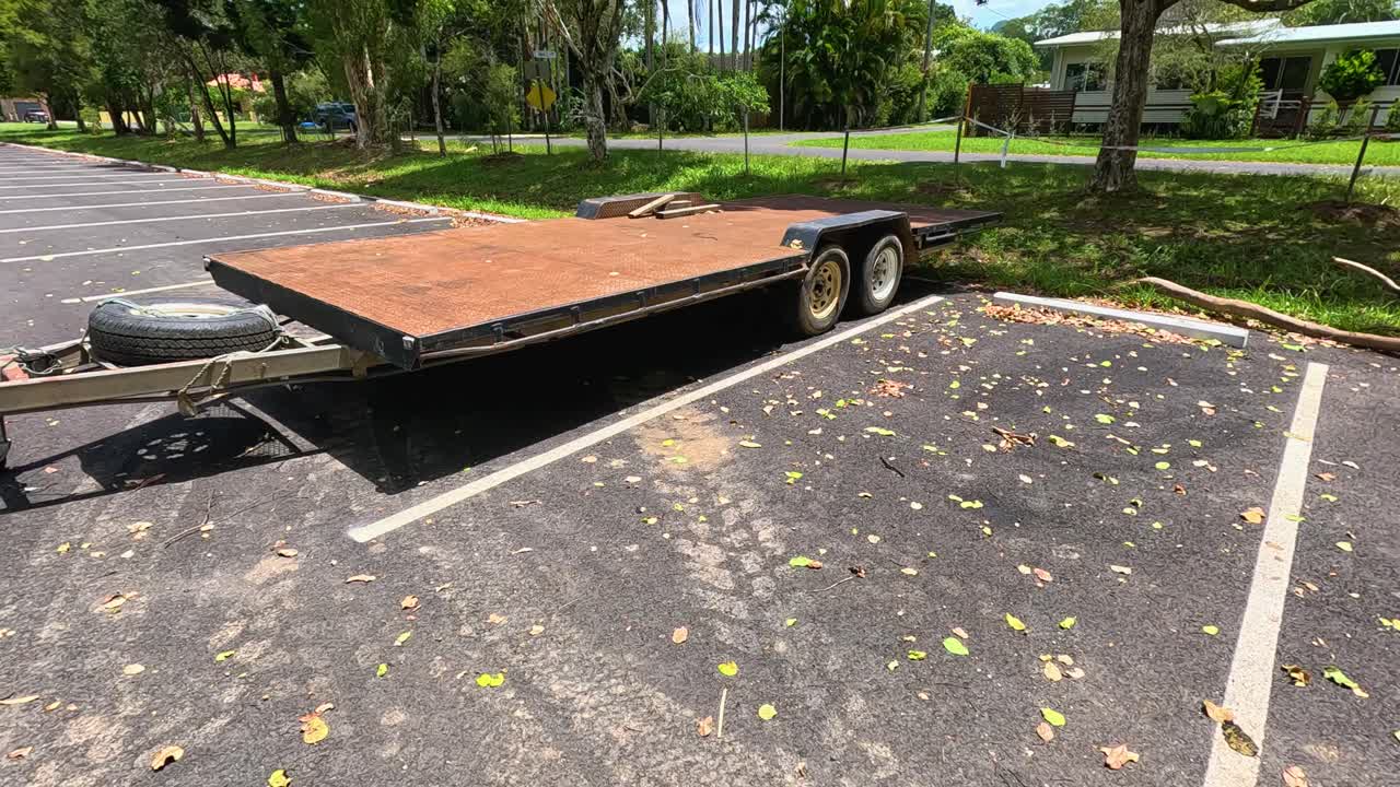 A stationary flatbed trailer sits in a sunlit parking lot, surrounded by trees and scattered leaves