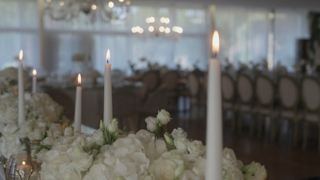 Close up of lit white candles and beautiful white floral arrangements on a luxury wedding reception table.