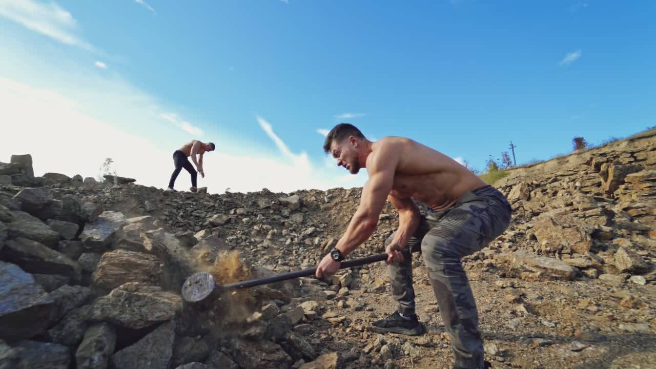 Athletic men doing hard exercises with stones. Shirtless sportsman breaks rocks with a sledgehammer in nature. Outdoor sport. Slow motion.