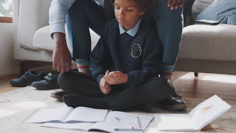 Niño-Preadolescente-De-Raza-Mixta-Con-Uniforme-Escolar-Sentado-En-El-Suelo-En-Casa-Haciendo-Su-Tarea-Ayudado-Por-Su-Padre,-Sentado-Detrás-De-él-En-El-Sofá,-Tiro-Inclinado-Con-La-Mano