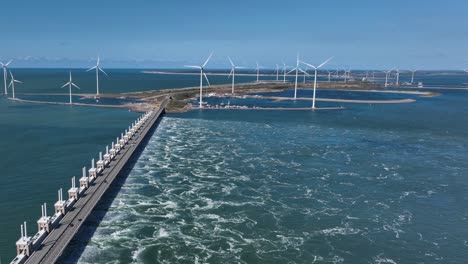 aerial flyover storm surge barrier dam at delta works in netherlands with rotating offshore wind turbines in background - cars crossing zeeland bridge