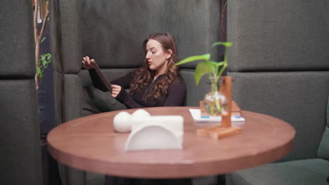 young woman with wavy hair sits on cushioned seat holding book, surrounded by modern decoration, visible brick wall in background, white decorative balls and small plant vase arranged on wooden table