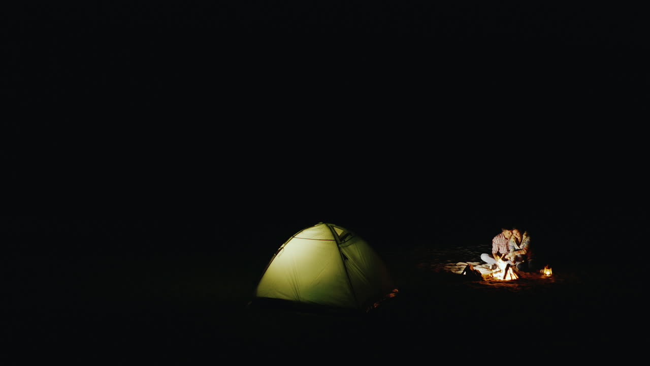 모닥불 주변에서 편안한 저녁을 보내는 젊은 부부와 위에서 텐트를 볼 수 있습니다.