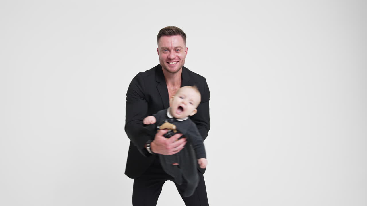 Happy smiling Caucasian dad waving his infant kid. Man in black suit playing with his lovely son. White backdrop.