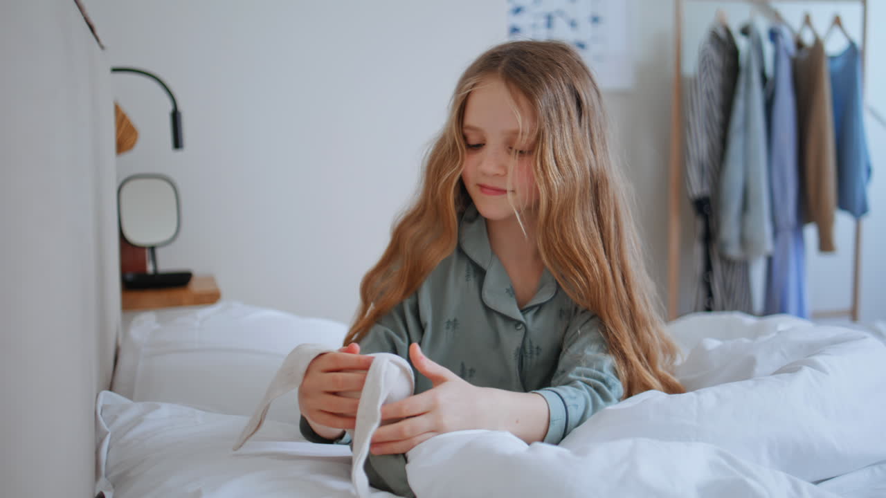 Bambina spensierata seduta sul letto che gioca con un peluche, primo piano. Bambina che si tocca i capelli