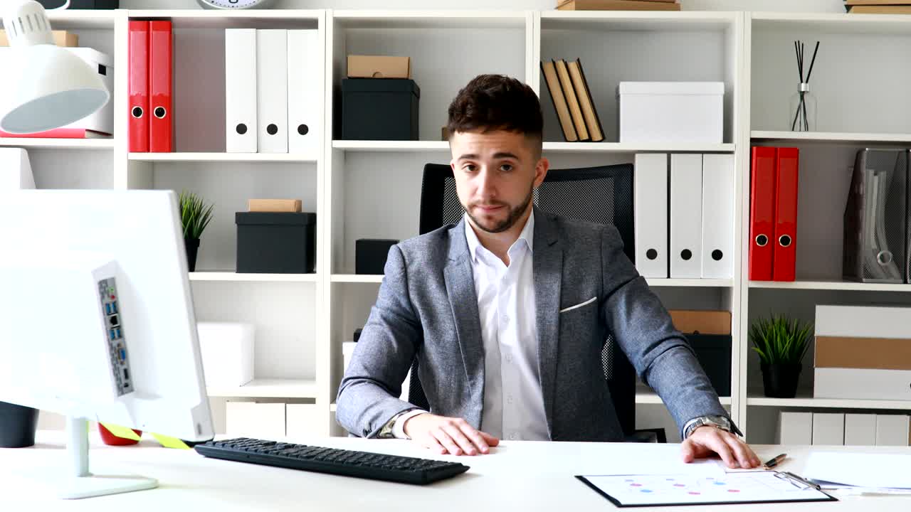 бизнесмен в сером пиджаке сидит за столом в белом офисе и покачивает головой негативно