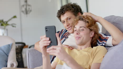 happy diverse gay male couple sitting at sofa using smartphone at home, slow motion