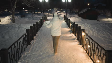 dancer wearing khaki pants white puffer coat and headphones dances on snow covered pavement lined with glowing lights and lampposts under night sky swaying arms and stepping with rhythmic joy