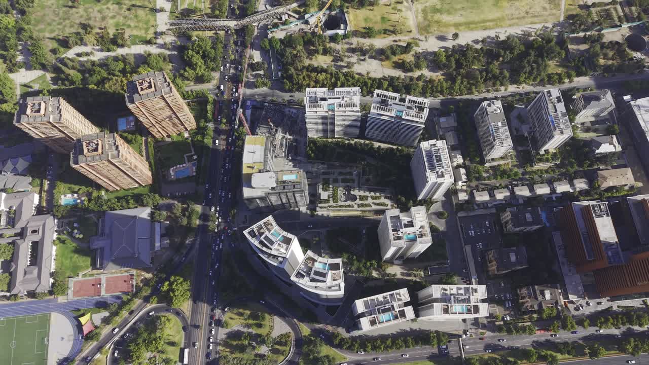 Vista aérea superior de um movimentado cruzamento urbano em Santiago, Chile, com prédios de apartamentos modernos com piscinas na cobertura, parques verdes exuberantes e intenso fluxo de tráfego