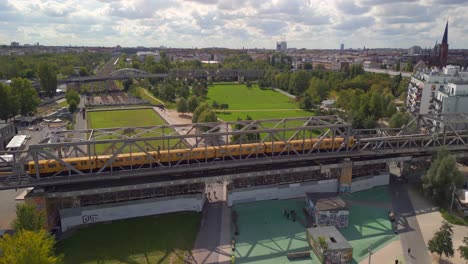 Vista-Aérea-Suave-Sujeto-De-Vuelo-Fuera-De-La-Vista-Drone-De-Un-Tren-Amarillo-U-bahn-En-Un-Puente-Ferroviario-De-Acero-En-El-Parque-En-Gleisdreieck-Berlín-Alemania-En-El-Día-De-Verano-De-2022