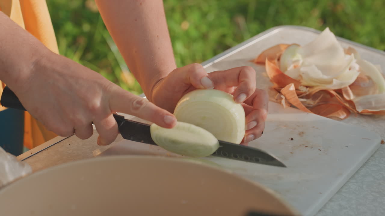 Woman slicing onion on board at campsite with focused hand technique and knife work, morning sunlight through trees, peelings piled beside board and prep bowl ready for skillet, intimate outdoor