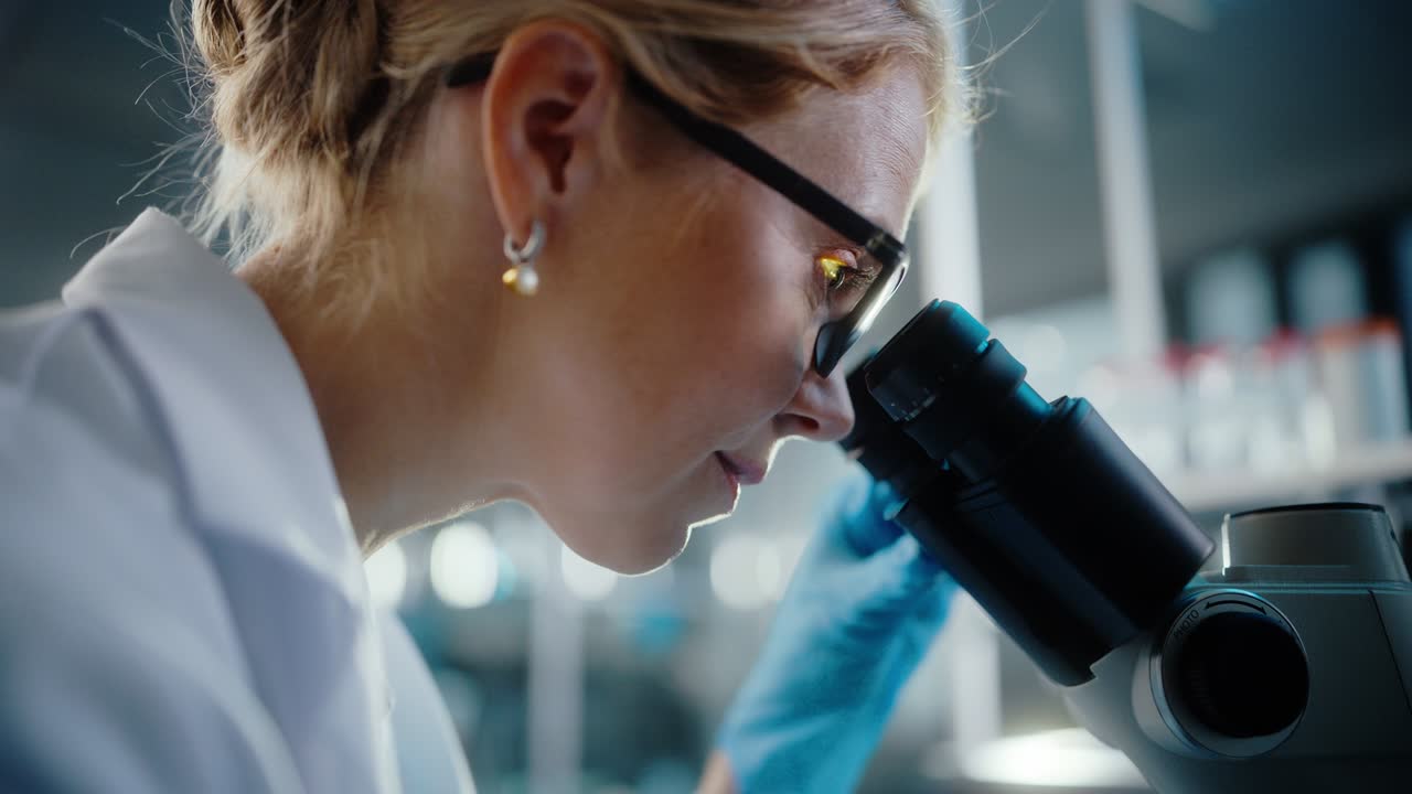In Bright Medical Science Laboratory: Beautiful Microbiologist Wearing Glasses Looks Under Microscope Analyzing Sample. Brilliant Scientist working with High-Tech Equipment. Close-up Shot