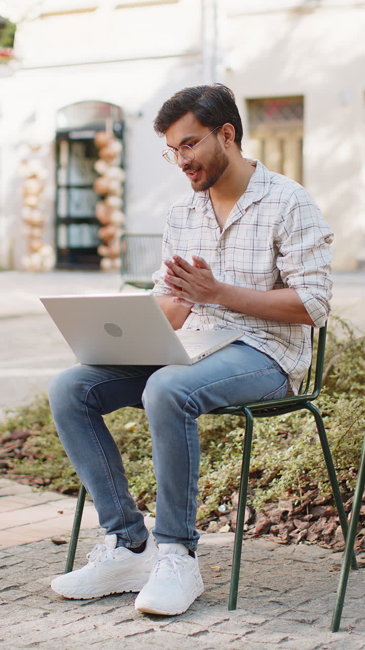 Giovane uomo indiano freelance seduto su una sedia che guarda la fotocamera del laptop mentre effettua una videochiamata all'aperto