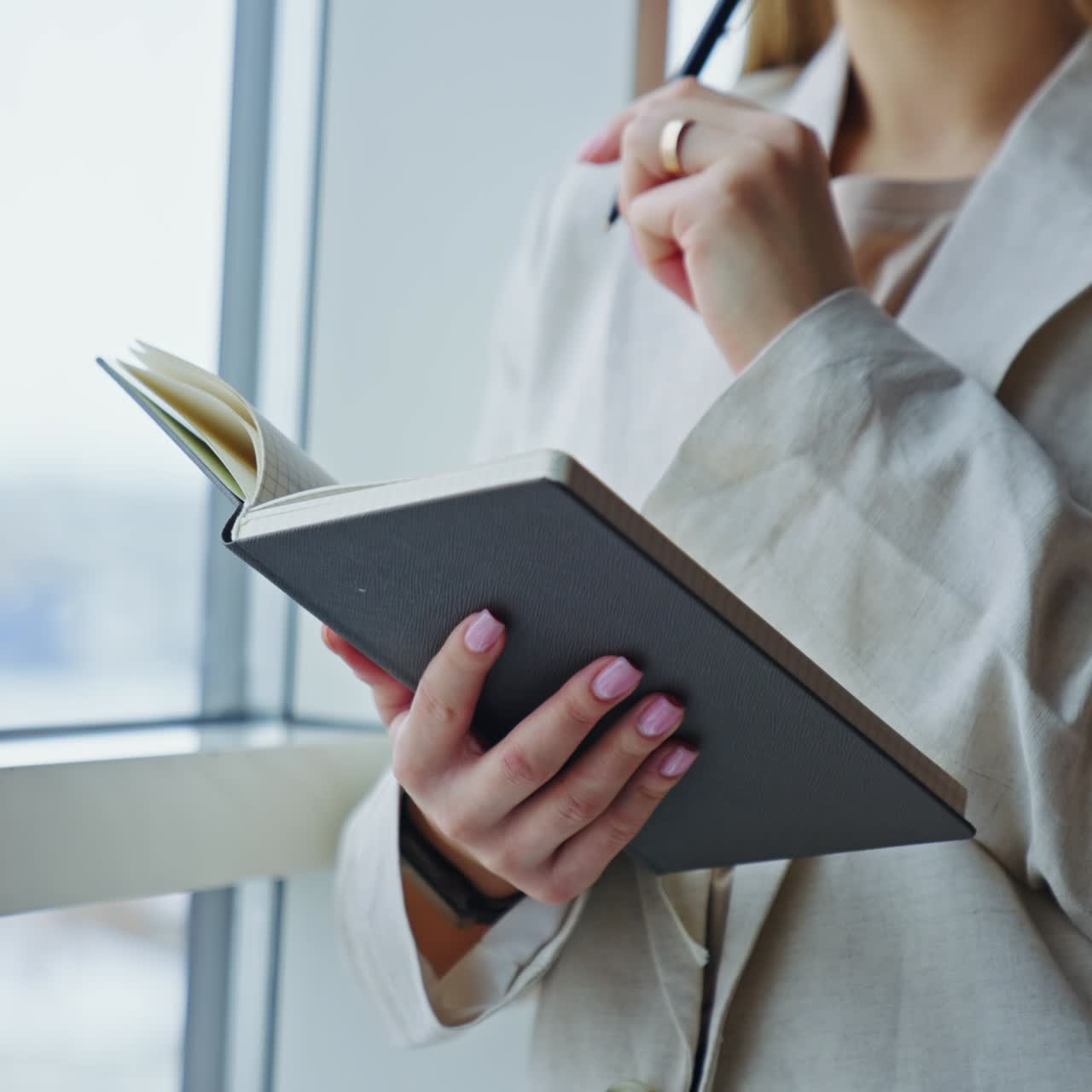 Woman in light jacket standing at the big window. Lady holding a notebook, writing something down there and closing it. Close up