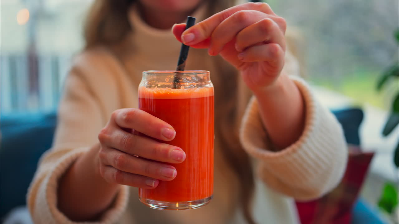 Close up of a woman mixing an orange and carrot juice in a glass with a black straw on a wooden table