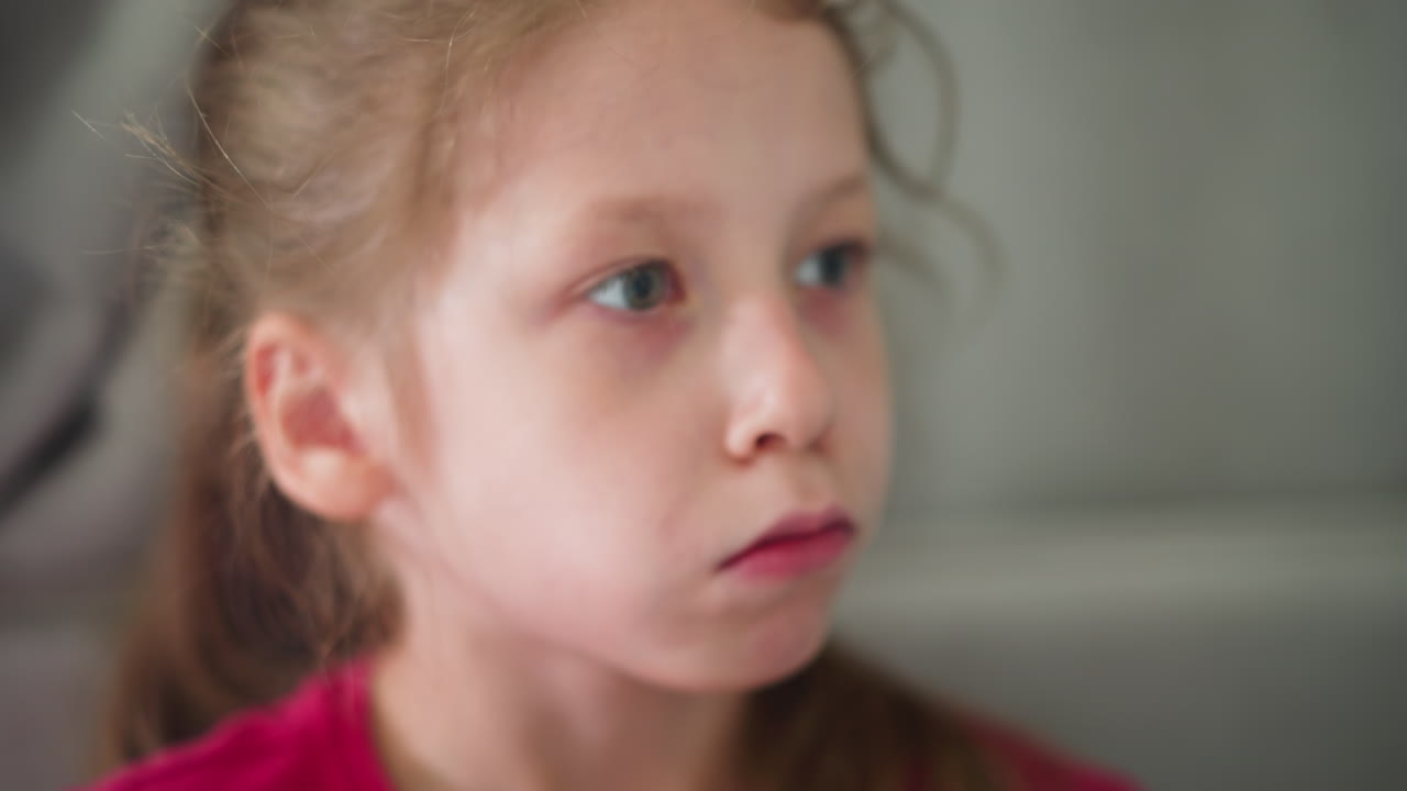 Close up of little girl wearing pink shirt eating snacks while looking curiously at someone in background indoors showing childhood innocence curiosity