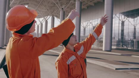 Dos-Trabajadores-Con-Uniforme-Naranja-Y-Casco-Apuntando-Al-Edificio-Y-Examinando-El-Objeto.-Camara-Lenta