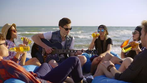 group of friends drinking cocktails and beer and doing cheers sitting on easychairs on the beach and listening to a friend playing guitar on a summer evening. shot in 4k.