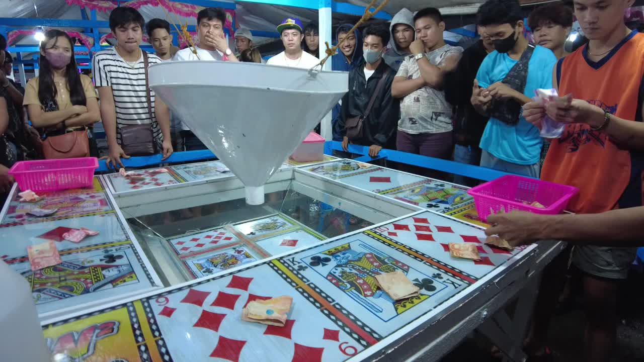 Closeup shot of tourists placing their money to place bets on the cards of their choice. The game attendant waits for the players to finish betting before signaling to toss the balls in the funnel.