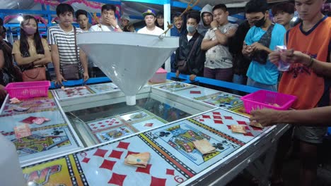 closeup shot of tourists placing their money to place bets on the cards of their choice. the game attendant waits for the players to finish betting before signaling to toss the balls in the funnel.